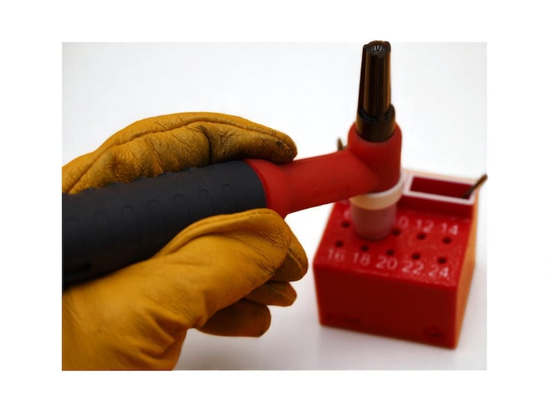 May include: A welding torch with a black handle and red nozzle is held by a person wearing yellow leather gloves. The torch is placed on a red block with numbered holes, likely for welding tips.