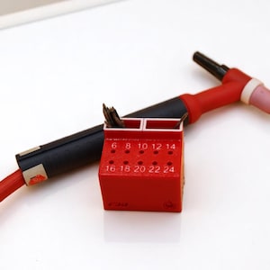 May include: A red and black welding torch with a red holder containing welding tips. The holder has numbers from 6 to 24 printed on it. The torch is on a white surface.