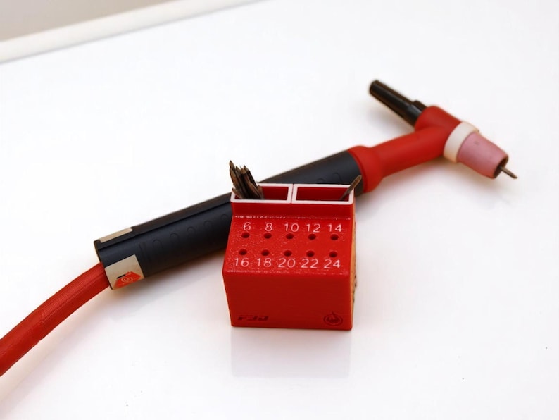 May include: A red and black welding torch with a red holder containing welding tips. The holder has numbers from 6 to 24 printed on it. The torch is on a white surface.