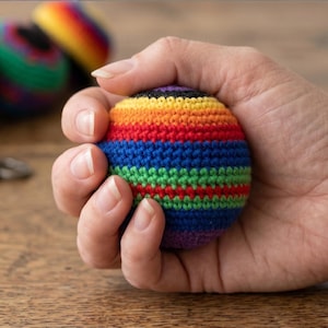 May include: A hand holding a small, round, crocheted ball with horizontal stripes in rainbow colors. Other colorful balls are blurred in the background, along with a vintage key on a wooden surface. The ball is approximately 2.5 inches in diameter.