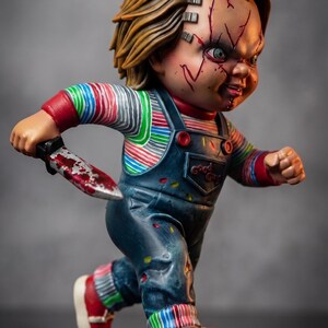 May include: A Chucky figure in a running pose, holding a bloody knife. The figure has blonde hair, facial scars, and is wearing a striped shirt, blue overalls, and red shoes. The figure is a collectible item.