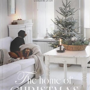 May include: A magazine cover titled Jeanne d'Arc Living's Special Christmas edition 2026 and The home of CHRISTMAS. The scene features a white sofa with two dogs, a white wooden table, a small Christmas tree, and a candle in a dark metal holder.