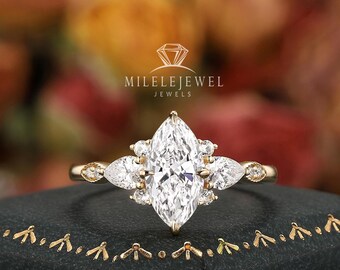May include: A marquise-cut diamond engagement ring with side stones, set in a gold band. The ring is displayed on a black jewelry box with gold trim. The ring features the text "MILELEJEWEL JEWELS".