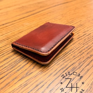 May include: A handcrafted, reddish-brown leather wallet with black stitching. The wallet is rectangular and appears to be folded, with visible layers. The logo "ZILCH RANCH" is stamped on the wooden surface beneath the wallet.