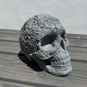 Può includere: Una scultura di teschio grigia dettagliata con intricati disegni floreali e a volute. Il teschio è realizzato in un materiale liscio e opaco e ha un aspetto realistico.