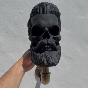 Puede incluir: Escultura de calavera negra con barba y pelo largos, montada en una tubería. La calavera tiene un diseño detallado y está hecha de un material liso y mate.