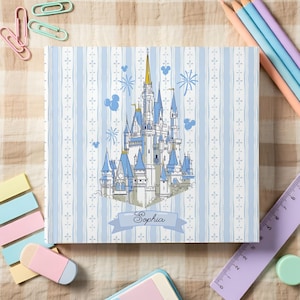 May include: A square, light blue notebook with a castle illustration and the name "Sophia" on a banner. The notebook has a striped pattern and is surrounded by pencils, a ruler, and stationery.