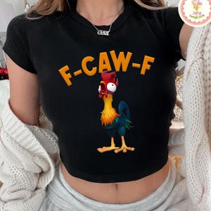 以下が含まれることがあります： オレンジ色の「F-CAW-F」の文字と漫画のニワトリのイラストが描かれた黒いクロップドトップス。ニワトリは赤、黄、青、緑色です。このトップスは半袖です。