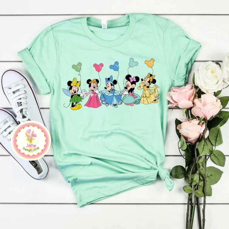 May include: A light green t-shirt featuring a graphic of cartoon characters holding balloons. The characters are dressed in colorful outfits. White sneakers and a floral design are visible on the left. A bouquet of pink and white roses is on the right.