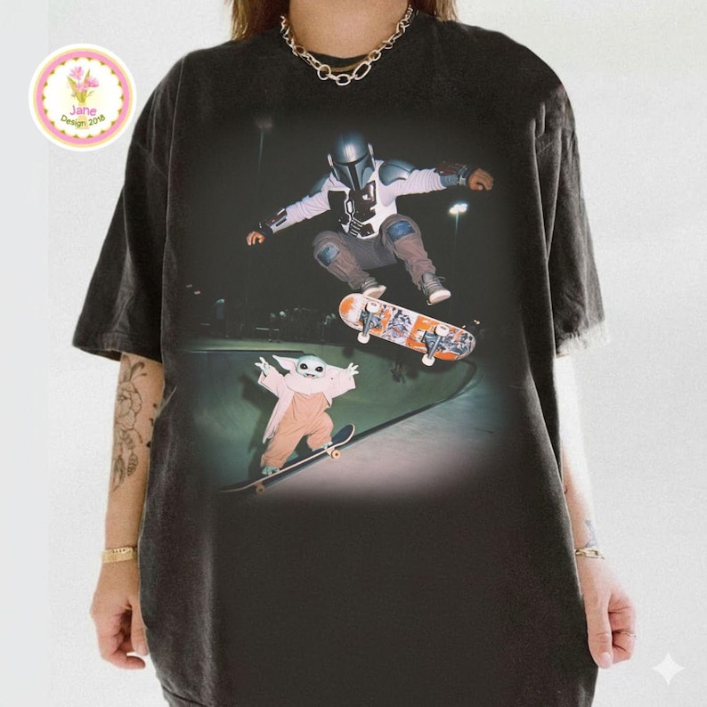 May include: Dark gray t-shirt featuring a graphic of a character in a helmet and another character, both skateboarding in a skate park. The skateboard has an orange and white design. The t-shirt has a distressed look.
