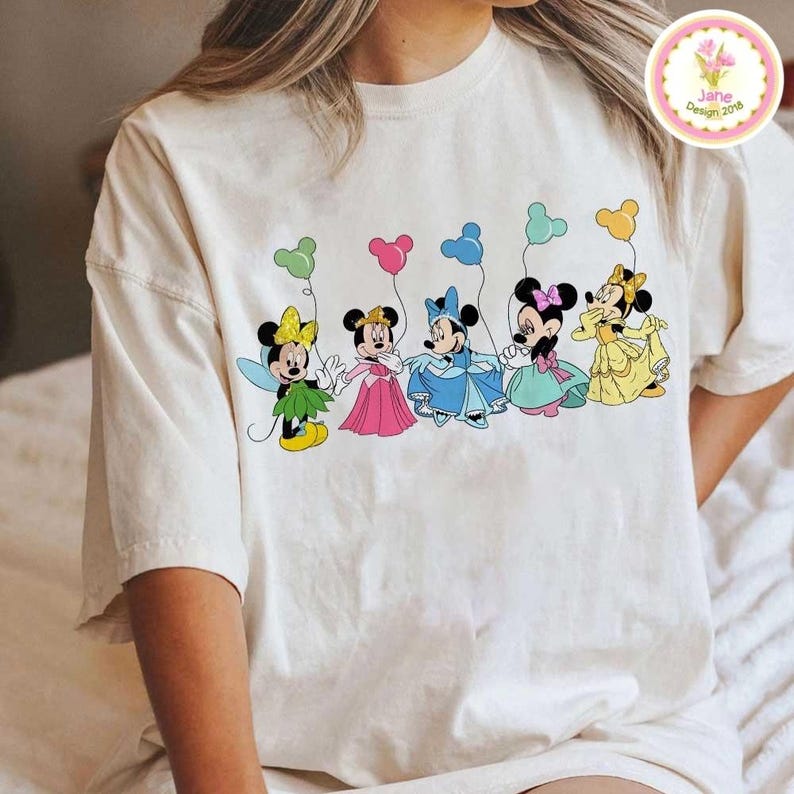 May include: Cream-colored t-shirt featuring a colorful graphic of five Minnie Mouse characters in various outfits, each holding a balloon. The balloons are in shades of green, pink, blue, teal, and yellow. The shirt has a relaxed fit and short sleeves.
