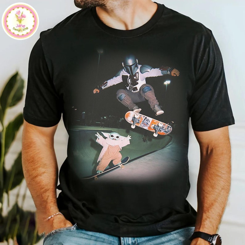 May include: Black t-shirt featuring a graphic of a skateboarder in a helmet and armor, mid-air, with a small figure on a skateboard below. The graphic is set in a skate park. The shirt has a crew neck and short sleeves.