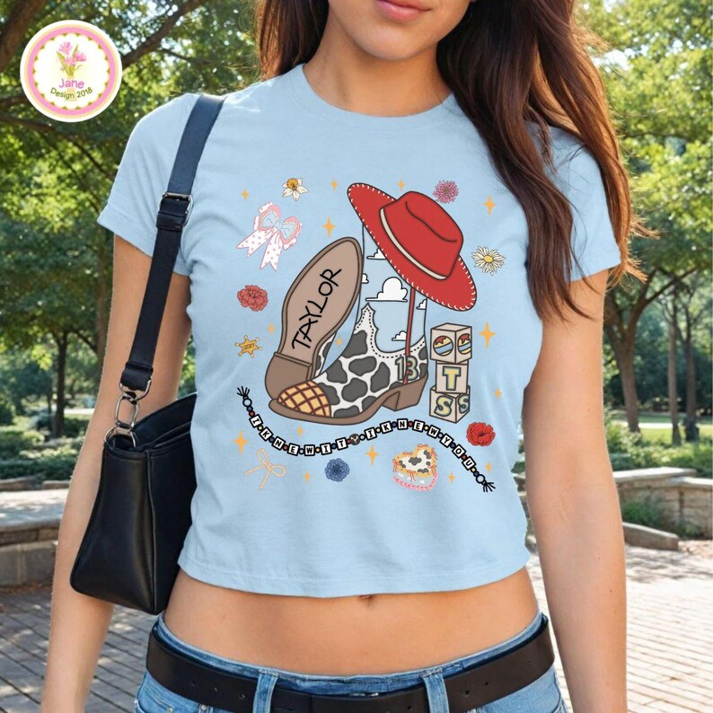 May include: A light blue short-sleeved t-shirt featuring a graphic print of a cow-print cowboy boot with the name Taylor, a red cowboy hat, stacked blocks with the letters T, S, and a beaded string with the text HE-NEW-IT-I-KNEW-IT-YOU.