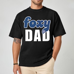 K&ouml;nnte beinhalten: Schwarzes T-Shirt mit dem Schriftzug "foxy DAD" in wei&szlig;en Blockbuchstaben. Das Wort "foxy" ist in blauer Schrift mit einem Fuchsschwanz-Design gehalten. Das Shirt ist ein l&auml;ssiger Stil.