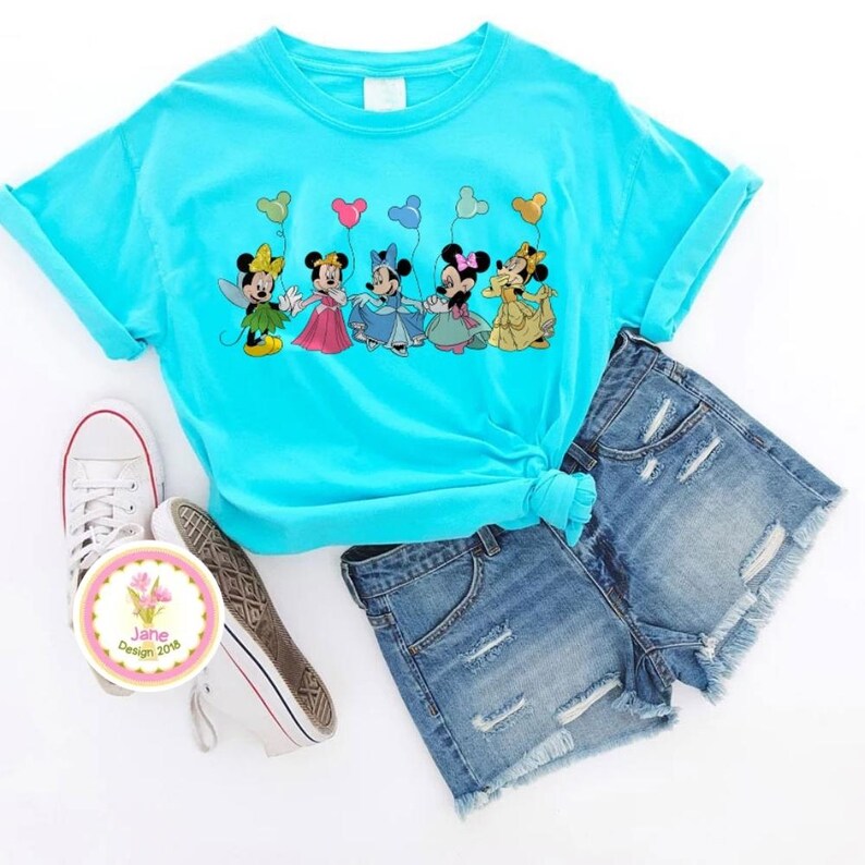 May include: A turquoise t-shirt featuring a graphic of five cartoon characters holding balloons. The shirt is paired with denim shorts and white sneakers. A circular sticker with floral design and text is also visible.