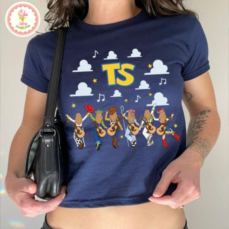 May include: A navy blue t-shirt featuring a graphic of six figures from the back, each holding a guitar. Above them are white clouds, yellow stars, musical notes, and the yellow text TS. The figures wear various outfits including hats and boots.