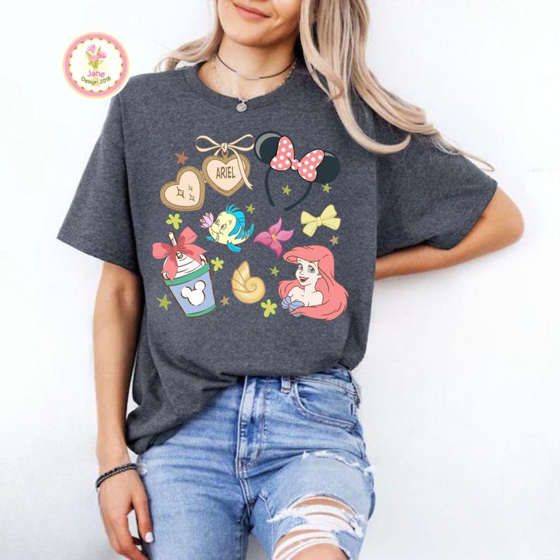 Op de afbeelding: Donkergrijs T-shirt met kleurrijke cartoonafbeeldingen van Ariel, een zeeschelp, een drankje en andere thema-elementen. Het woord "ARIEL" staat op een hartvormige koek. Het shirt heeft een ronde hals en korte mouwen.