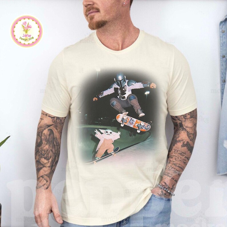 May include: Cream-colored t-shirt featuring a graphic of a character in a Mandalorian helmet and a Grogu character skateboarding. The graphic is set against a dark background. The shirt has short sleeves and a crew neck.