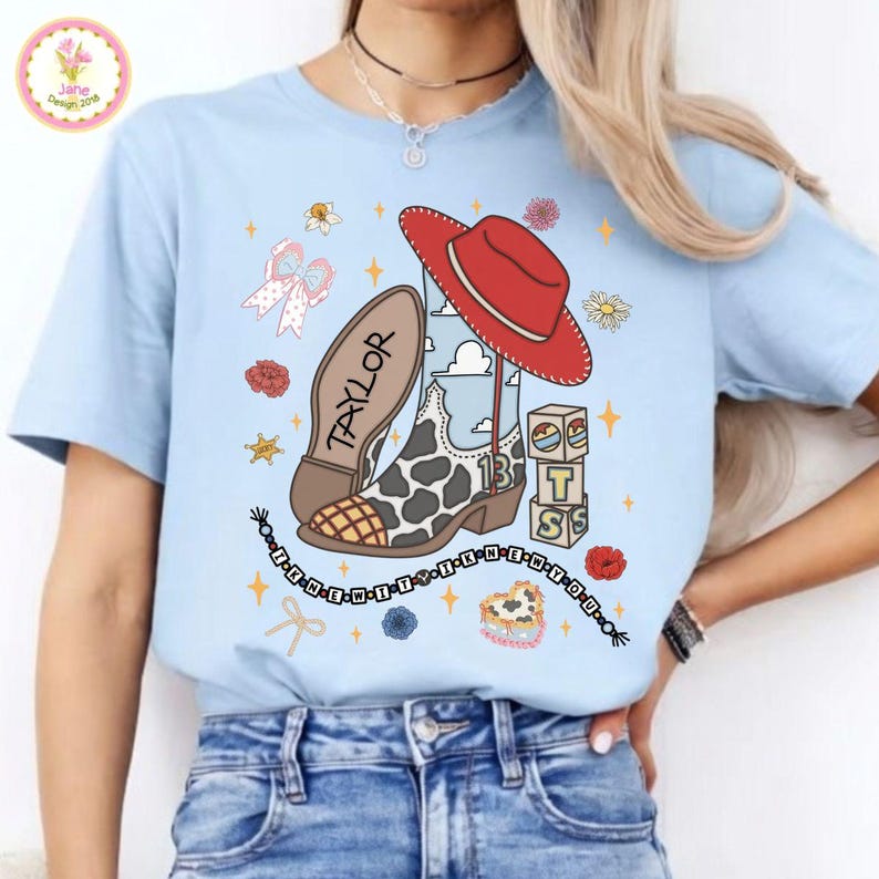 May include: A light blue t-shirt featuring a graphic print of a cow-print cowboy boot, a red cowboy hat, three stacked blocks with the letters T, S, and a sun icon, and a beaded string with the text I KNEW IT I KNEW YOU and the name TAYLOR.