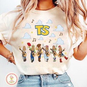 May include: A cream-colored t-shirt featuring a graphic print of six figures with long hair, each holding an acoustic guitar. Above them are blue clouds, yellow stars, musical notes, and the large yellow letters TS with a blue outline.