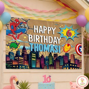 May include: A birthday banner featuring Spider-Man, the text "Happy Birthday Thomas!", and the number 6 in a comic book style. The banner also includes city skyline graphics and superhero emblems. Balloons and a cake are also visible.