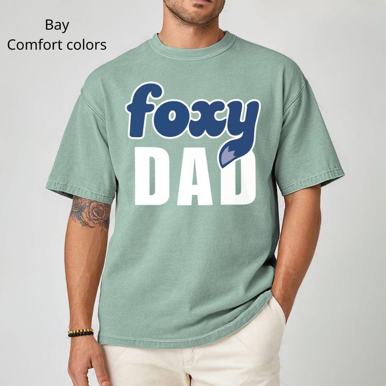 K&ouml;nnte beinhalten: Hellgr&uuml;nes T-Shirt mit den Worten "foxy DAD" in einem blau-wei&szlig;en Design. Das Wort "foxy" ist in blauer, kursiver Schrift geschrieben, und das Wort "DAD" ist fett und wei&szlig;. Das Shirt hat kurze &Auml;rmel.