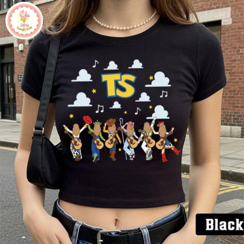 May include: A black short-sleeved crop top featuring a graphic print of six figures with guitars, white clouds, yellow stars, musical notes, and the yellow text TS. The shirt has a crew neckline and is worn with a black belt and a black shoulder bag.