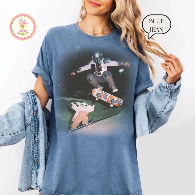 May include: Blue jean-colored t-shirt featuring a graphic of two Star Wars characters skateboarding in a skate park. The shirt has a relaxed fit and short sleeves. The text "BLUE JEAN" is in a speech bubble. A logo is in the upper left corner.