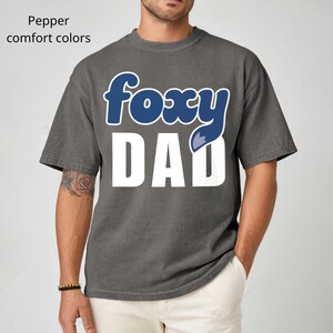 K&ouml;nnte beinhalten: Ein graues T-Shirt mit dem Text "foxy DAD" in Wei&szlig; und Blau. Das Wort "foxy" ist in einer blauen, kursiven Schriftart gehalten, und der Buchstabe "y" wird durch einen blauen Fuchsschwanz ersetzt. Das Shirt ist eine Komfortfarbe.