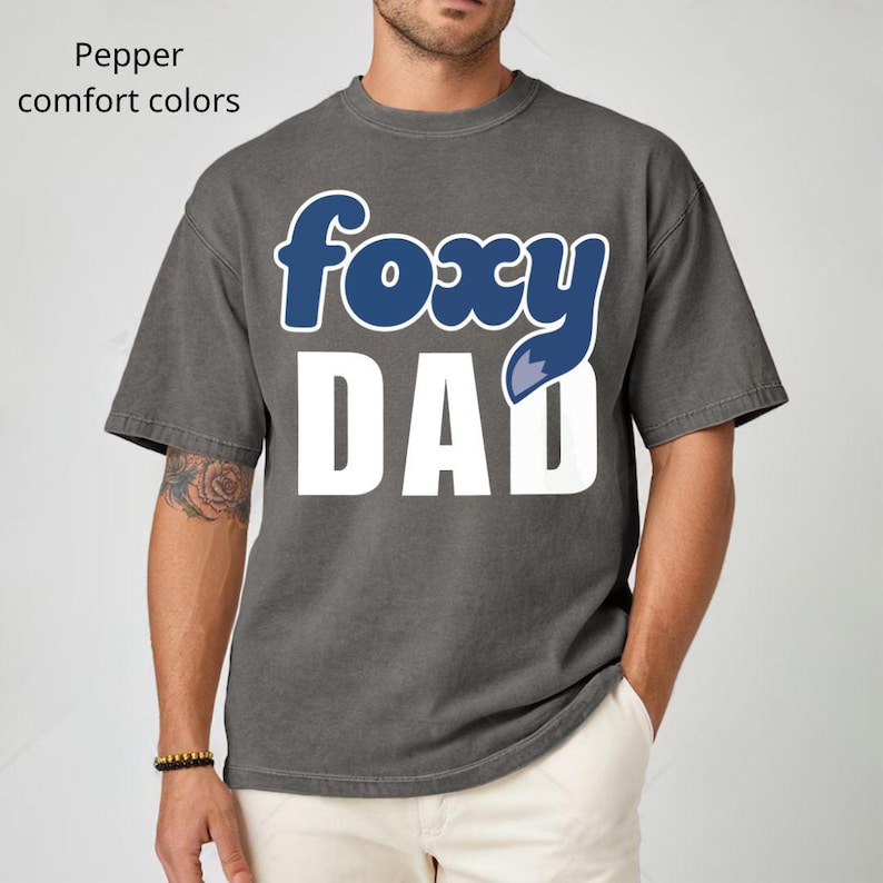 K&ouml;nnte beinhalten: Ein graues T-Shirt mit dem Text "foxy DAD" in Wei&szlig; und Blau. Das Wort "foxy" ist in einer blauen, kursiven Schriftart gehalten, und der Buchstabe "y" wird durch einen blauen Fuchsschwanz ersetzt. Das Shirt ist eine Komfortfarbe.