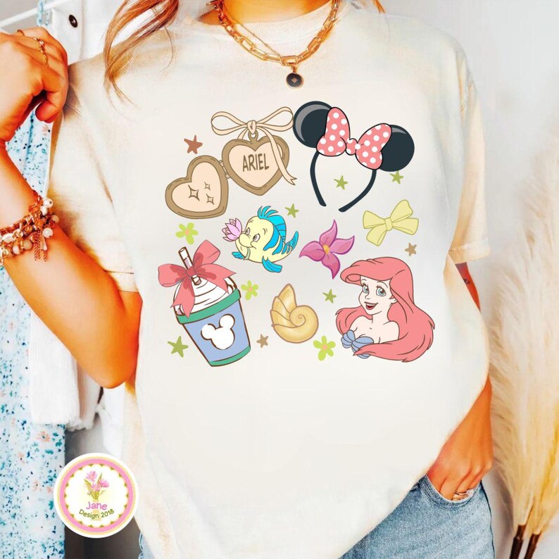 Op de afbeelding: Cr&egrave;mekleurig T-shirt met diverse cartoonafbeeldingen. Ontwerpen omvatten een Minnie Mouse strik hoofdband, Ariel de Kleine Zeemeermin, Botje, een zeeschelp en een Mickey Mouse drankje. Het woord "ARIEL" staat op een hartvormige hanger.