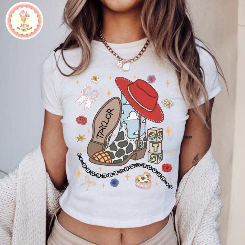May include: A white short-sleeved t-shirt featuring a graphic print of a cow-print cowboy boot, a red cowboy hat, alphabet blocks spelling TS, and a beaded necklace with the text I KNEW IT IN MY YOU. The boot has the name TAYLOR printed on it.