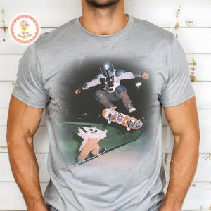 May include: Gray t-shirt featuring a graphic of a character in a Mandalorian helmet and another character, both skateboarding. The skateboarders are in a skate park. The shirt has a crew neck and short sleeves.