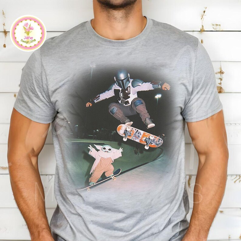 May include: Gray t-shirt featuring a graphic of a character in a Mandalorian helmet and another character, both skateboarding. The skateboarders are in a skate park. The shirt has a crew neck and short sleeves.