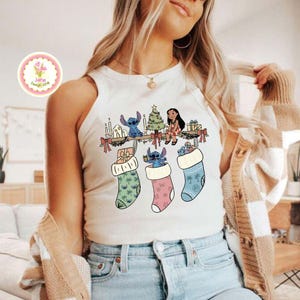 May include: A white tank top featuring a Christmas-themed graphic. The design includes cartoon characters, a Christmas tree, and stockings. The stockings are in pastel colors and filled with gifts. The overall theme is festive and whimsical.