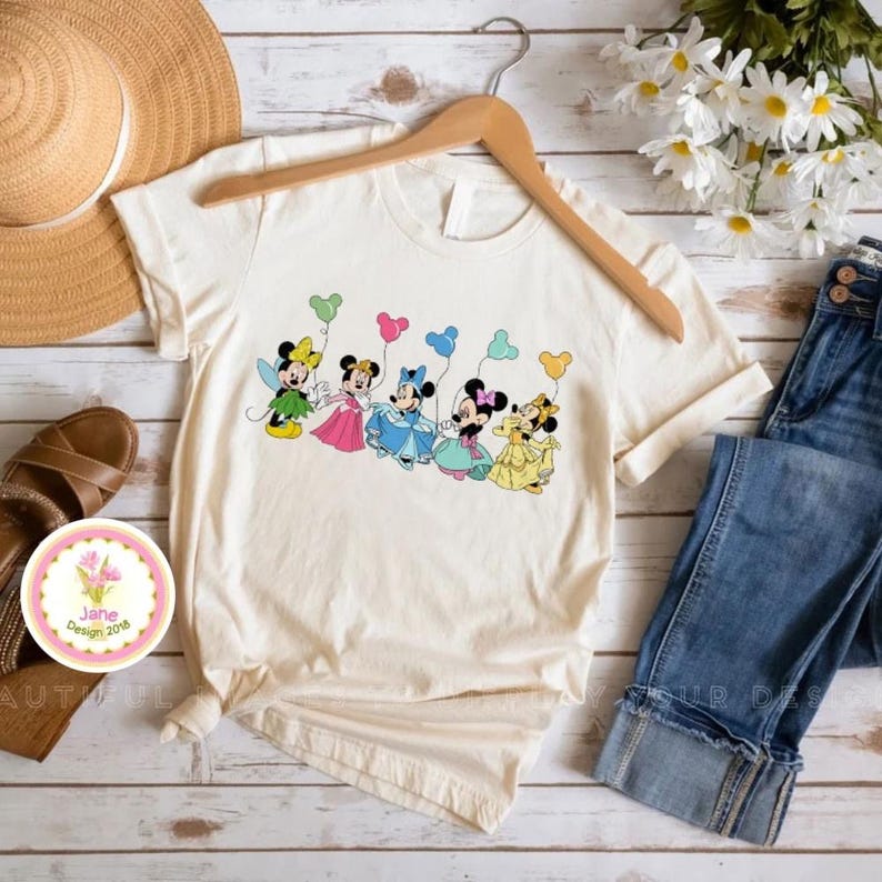 May include: Cream-colored t-shirt featuring a colorful graphic of six cartoon mice holding heart-shaped balloons. The mice are dressed in various outfits, including dresses and fairy attire. The shirt is hanging on a wooden hanger.