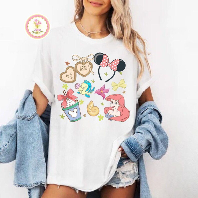 Op de afbeelding: Wit T-shirt met kleurrijke cartoonafbeeldingen van Disney-personages en accessoires. Inclusief een Minnie Mouse-haarband, een zeeschelp en de naam "Ariel" op een hartvormig label. Het shirt heeft een relaxte pasvorm.