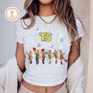 May include: A white short-sleeved t-shirt featuring a graphic print of six figures with long hair, each holding an acoustic guitar. Above the figures are blue clouds, musical notes, yellow stars, and the bold yellow and blue text TS.