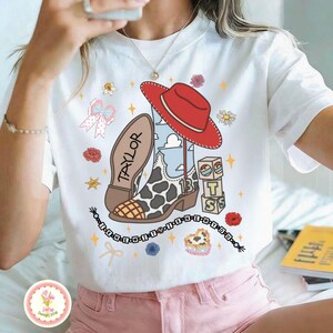 May include: A white t-shirt featuring a graphic print of a red cowboy hat, a cow-print western boot with the number 13, alphabet blocks spelling TS, a beaded string with the text I KNEW YOU WERE TROUBLE, and various floral and star icons.
