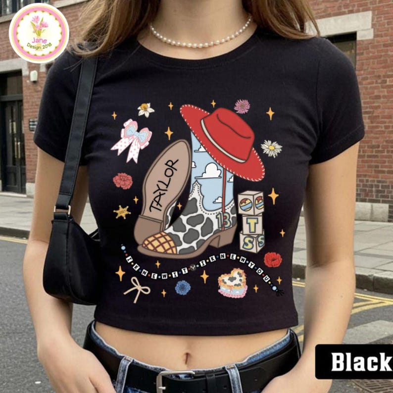 May include: A black short-sleeved t-shirt featuring a graphic print of a cowboy boot with a cow print pattern, a red hat, the name TAYLOR, the number 13, and blocks with the letters T, S, and a cloud design, surrounded by small stars and bows.