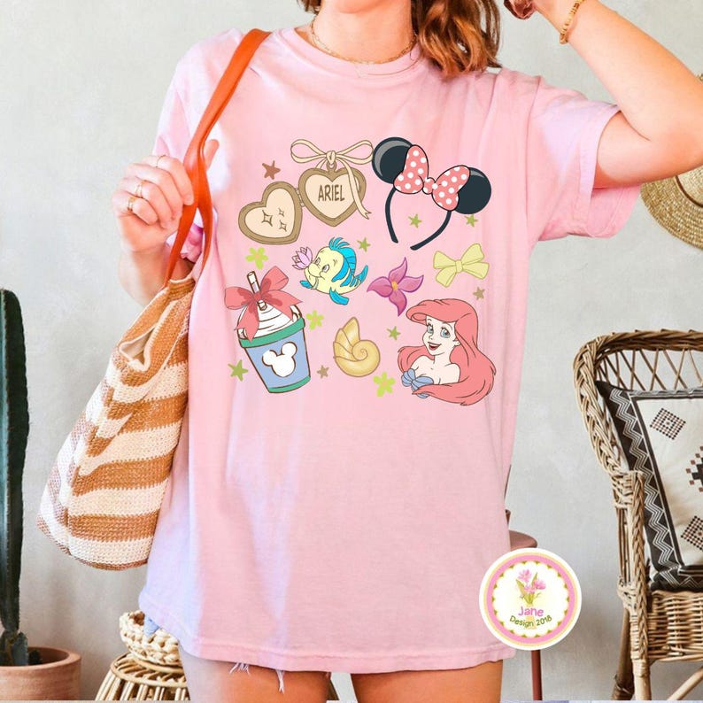 Op de afbeelding: Lichtroze T-shirt met cartoonafbeeldingen. Het ontwerp bevat een hartvormig label met de naam "ARIEL", zwarte muizenoren met een roze stippen strik en een cartoon zeemeermin. Andere afbeeldingen zijn een zeeschelp en een drankje.