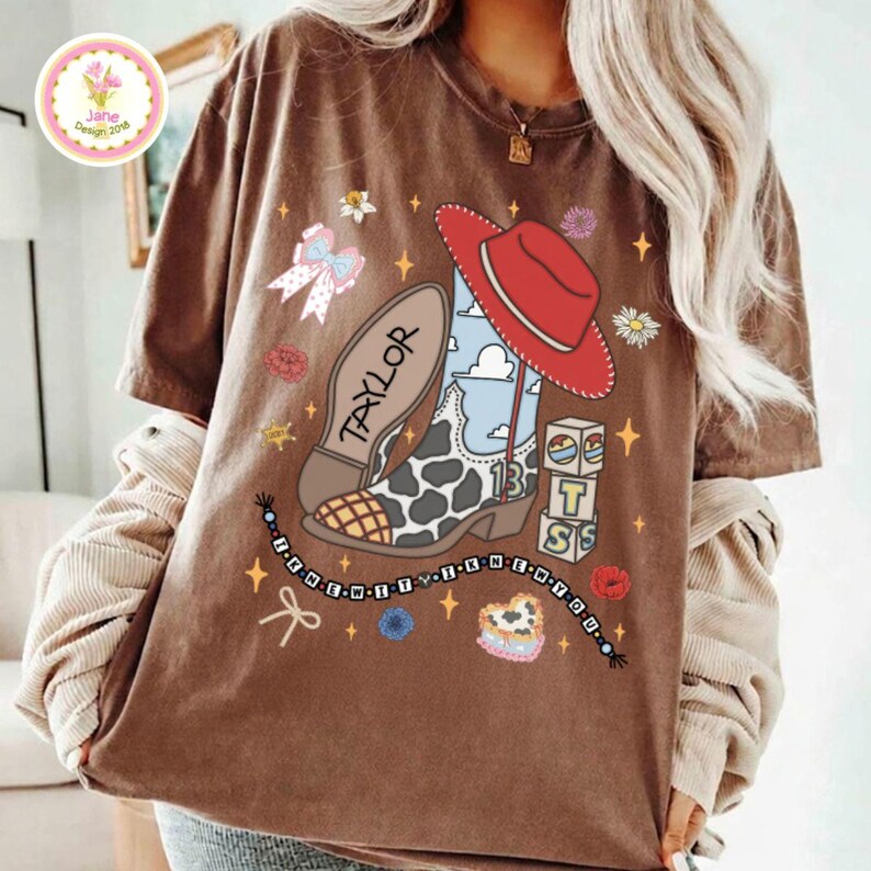 May include: A brown short-sleeve t-shirt featuring a graphic print of a cow-print cowboy boot with a red hat, the name Taylor, the number 13, stacked blocks, a bow, and flowers. The design includes a beaded string border and small star accents.