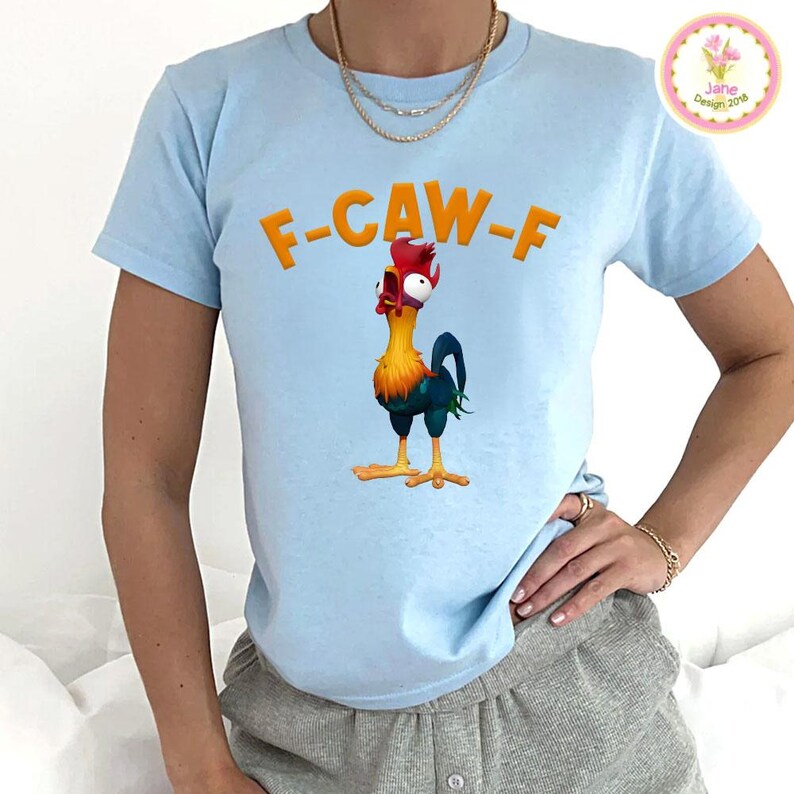 以下が含まれることがあります： オレンジ色の「F-CAW-F」の文字と、漫画のオンドリのイラストがプリントされた水色のTシャツ。オンドリは赤、黄、青、緑で描かれています。Tシャツはクルーネックで半袖です。