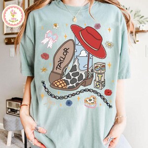 May include: A light green t-shirt featuring a graphic print of a red cowboy hat, a cow-print boot with the name TAYLOR, and blocks spelling TS. The design includes a beaded string with the text I KNEW IT I KNEW YOU, bows, and flowers.
