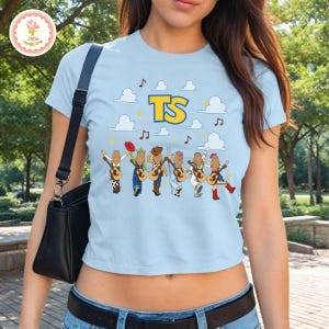 May include: A light blue short-sleeved t-shirt featuring a graphic of six figures with long hair, each holding an acoustic guitar. Above them are white clouds, yellow stars, musical notes, and the yellow text TS. A black shoulder bag is visible.