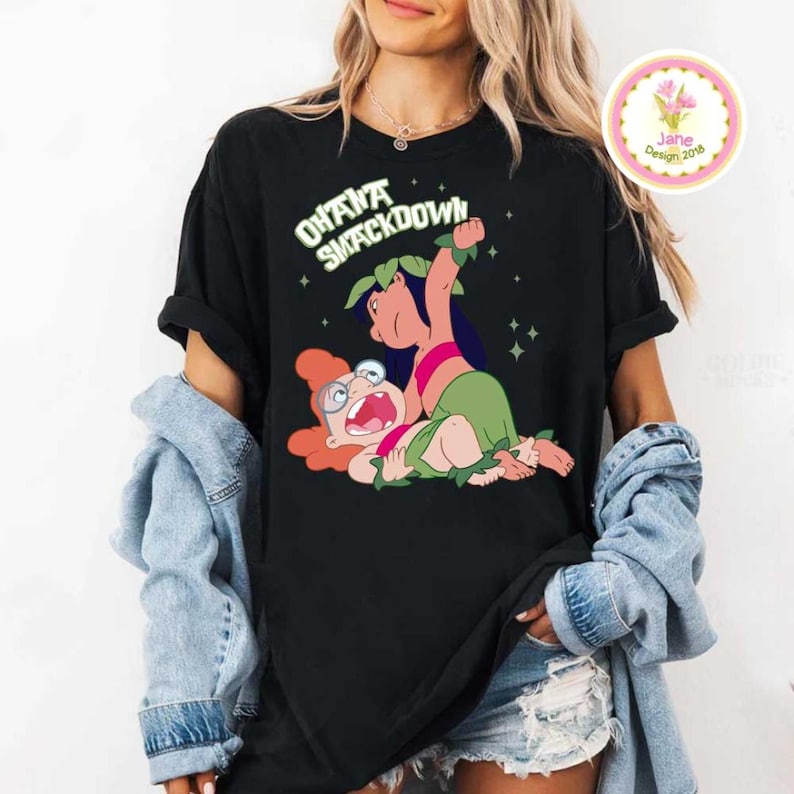 Może przedstawiać: Czarny t-shirt z kresk&oacute;wkową ilustracją dw&oacute;ch postaci w pozie wrestlingowej. Tekst "OHANA SMACKDOWN" jest wygięty nad postaciami. Małe, okrągłe logo znajduje się w prawym g&oacute;rnym rogu.
