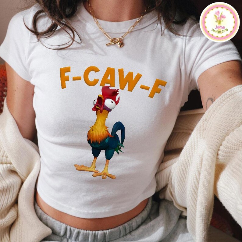 以下が含まれることがあります： オレンジ色の文字で「F-CAW-F」と書かれた白いクロップドトップス。テキストの下には、黄色、赤、青の羽を持つ漫画のオンドリが印刷されています。オンドリは口を開けて上を見上げています。