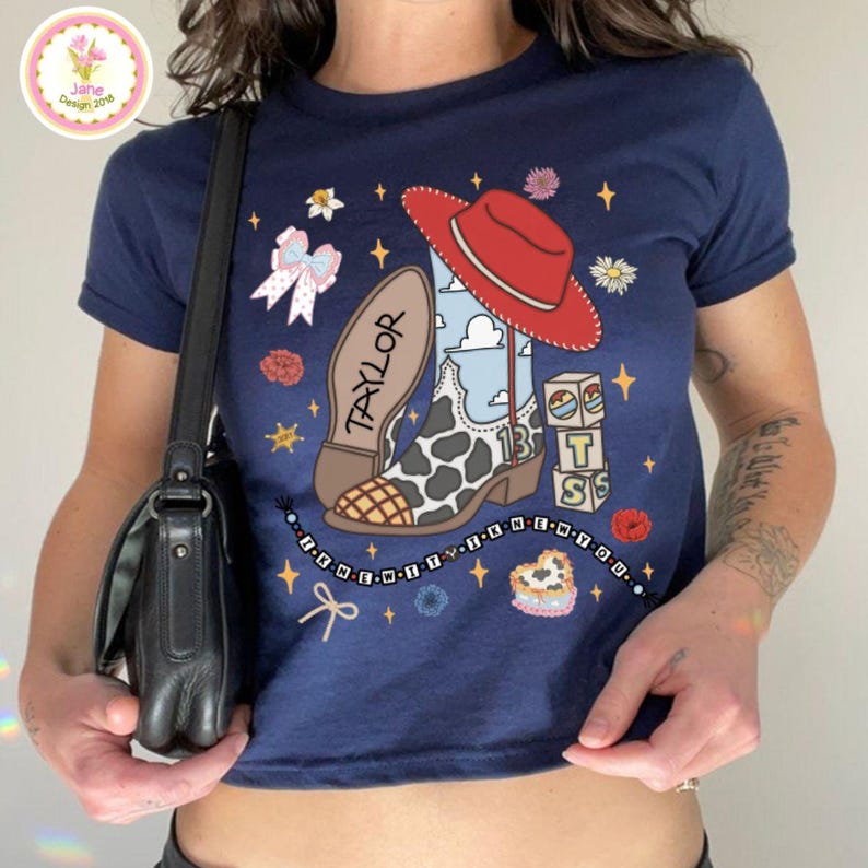May include: A navy blue t-shirt featuring a graphic print of a cowboy boot with a cow print pattern, a red hat, a pink bow, blocks with the letters T, S, and the number 13, and a beaded string with the text I KNEW IT I KNEW YOU.