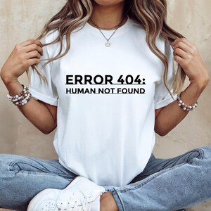 May include: White t-shirt with the text "ERROR 404: HUMAN NOT FOUND" in black. The shirt is a crew neck style and is being worn by someone wearing blue jeans and white sneakers.