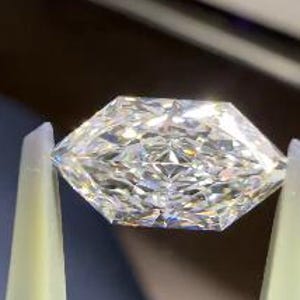 May include: A faceted, clear diamond held by tweezers. The diamond has a geometric shape with multiple facets that reflect light, creating a sparkling effect. The tweezers are light yellow and black.